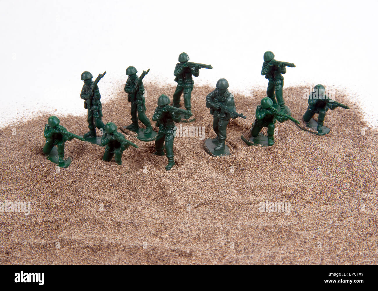 A group of little plastic green army men set up on a sandpile against a white background. Stock Photo