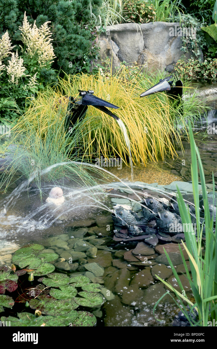 Water garden with funny bird fountains, frogs spouting streams in garden pond with plants lilypads ornamental grasses Stock Photo