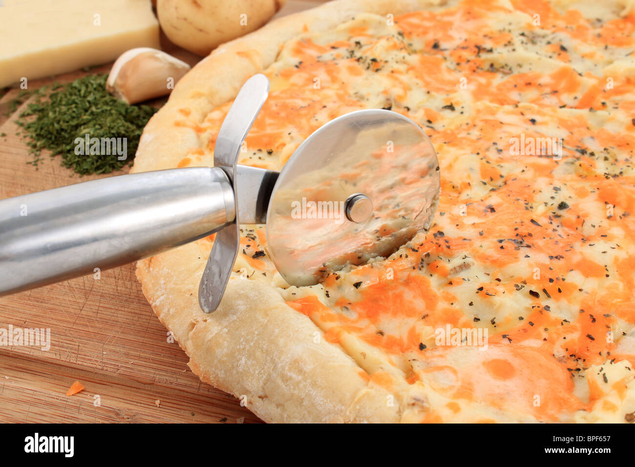 Silver cutter cutting through gourmet pizza made with fresh dough, potatoes, cheese, and garlic with seasoning Stock Photo