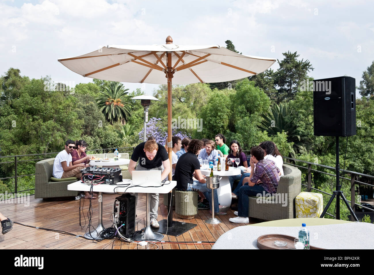 deejay hovers over Macbook as groups of attractive young Mexicans relax with drinks at trendy upscale rooftop bar Hotel Condesa Stock Photo