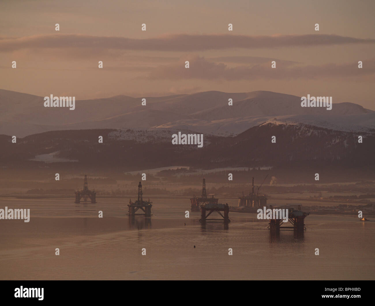 Oil rigs in a Scottish Harbour, are overshadowed by snow covered mountains as the sun sets. Stock Photo