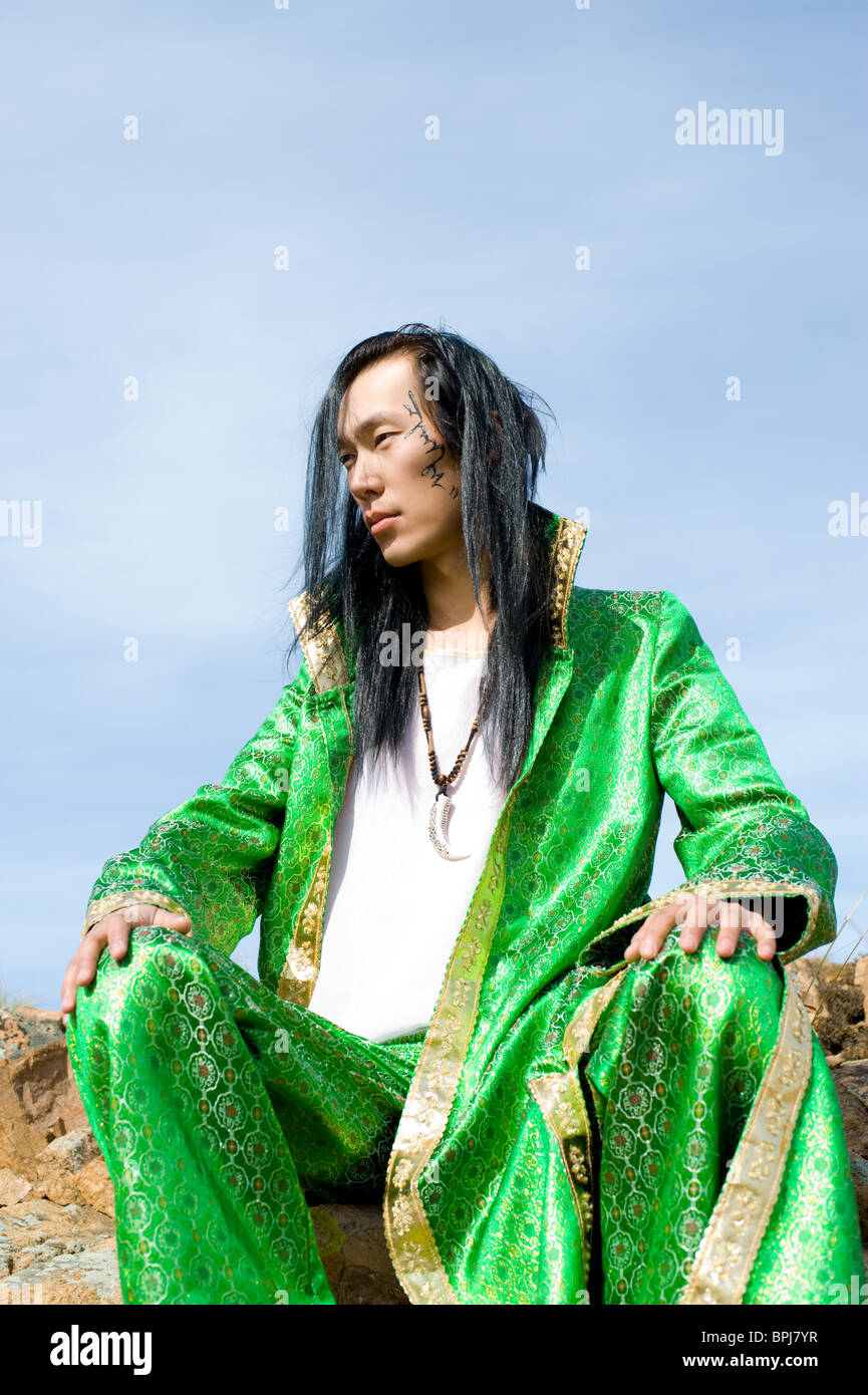 young asian man in green Mongolian suit sits on rock, his name Chinghis is written in ancient Mongolian on his cheek Stock Photo