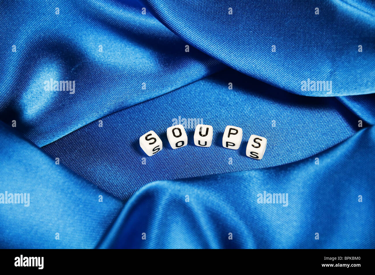 Royal blue satin background with rich folds and wrinkles for texture is the word Soups in black and white cube lettering series. Stock Photo