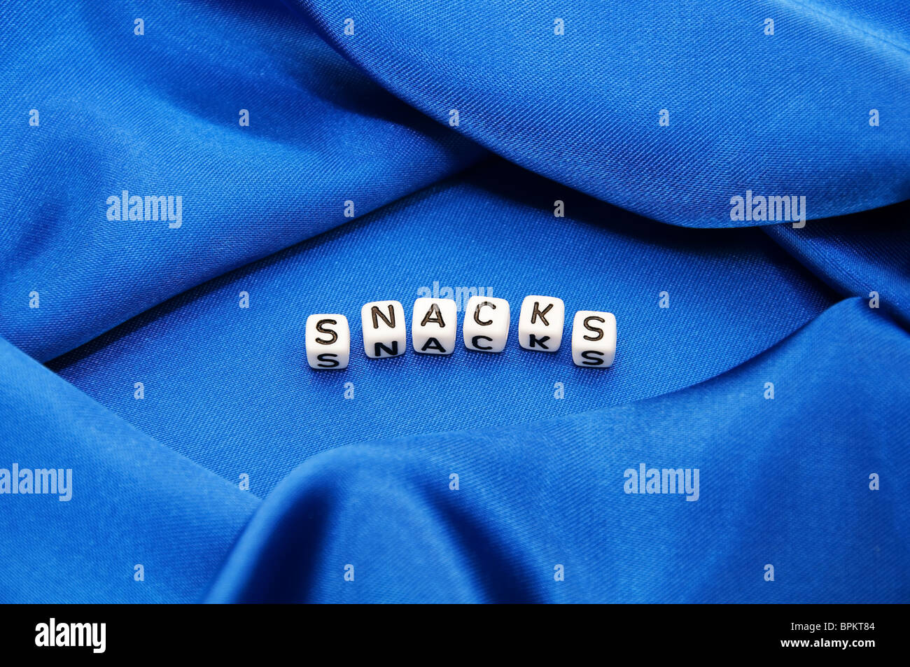 Royal blue satin background with rich folds and wrinkles for texture is the word snacks in  cube lettering in this series. Stock Photo