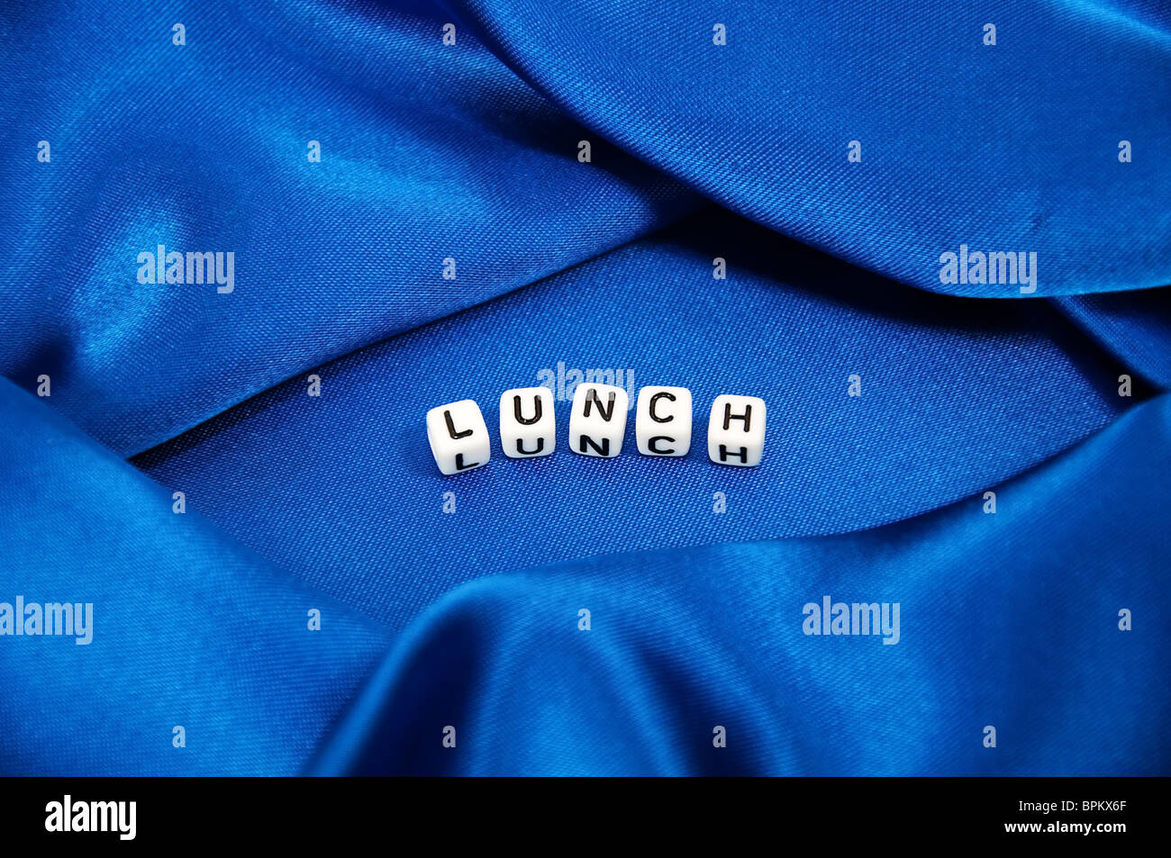 Royal blue satin background with rich folds and wrinkles for texture is the word lunch in black and white cube lettering series. Stock Photo