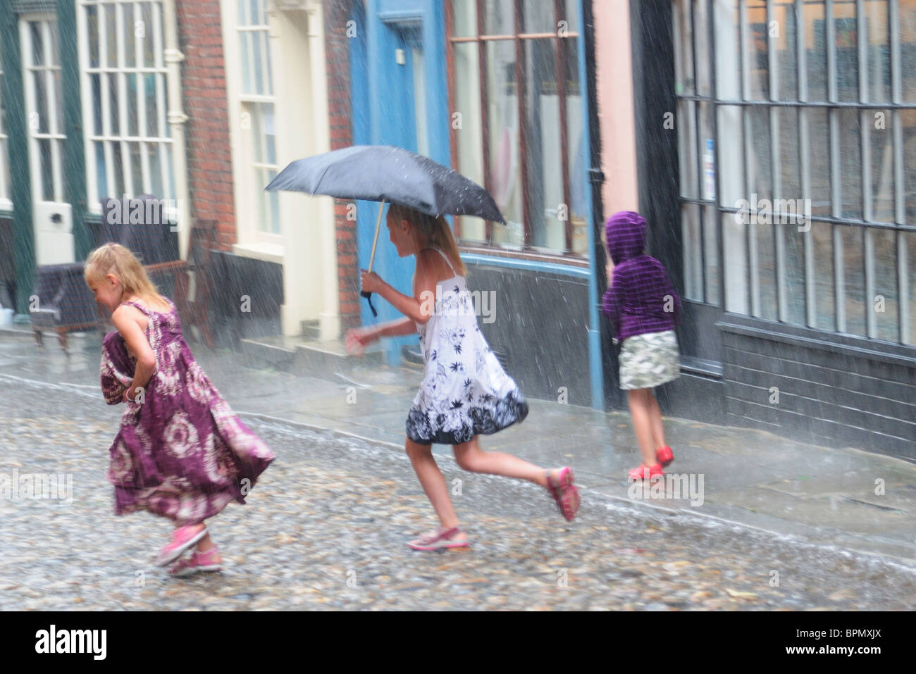 Three young girls running in the summer rain in cobbled street. Stock Photo