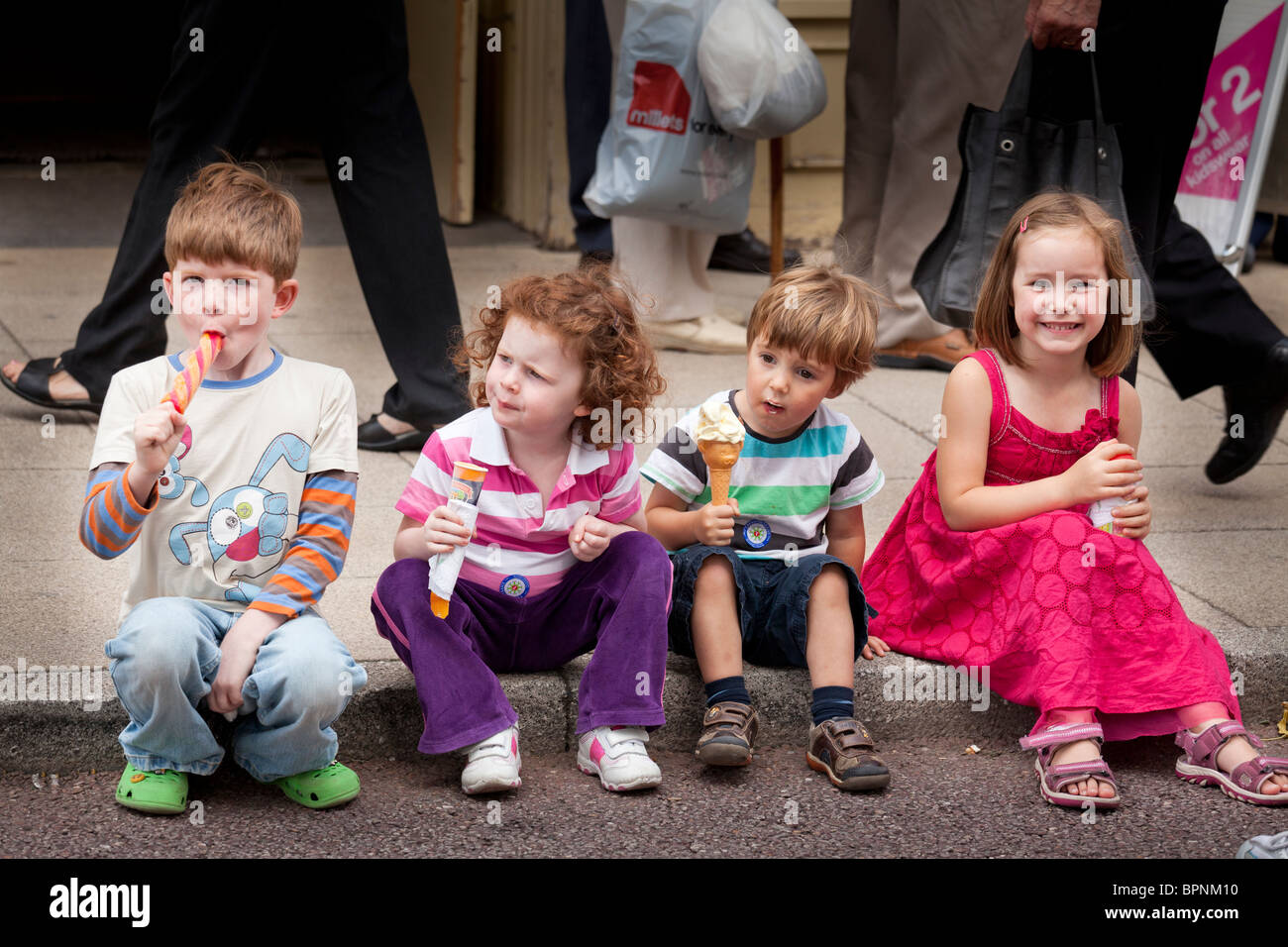 Four young children sitting on pavement kerb eating ice creams. Stock Photo