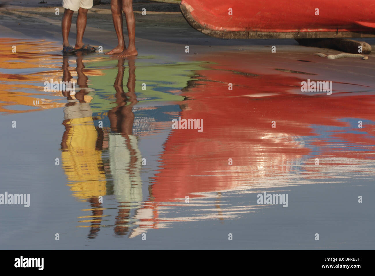 Inhabitants of Goa standing by a boat are reflected in the water on the shore. Stock Photo