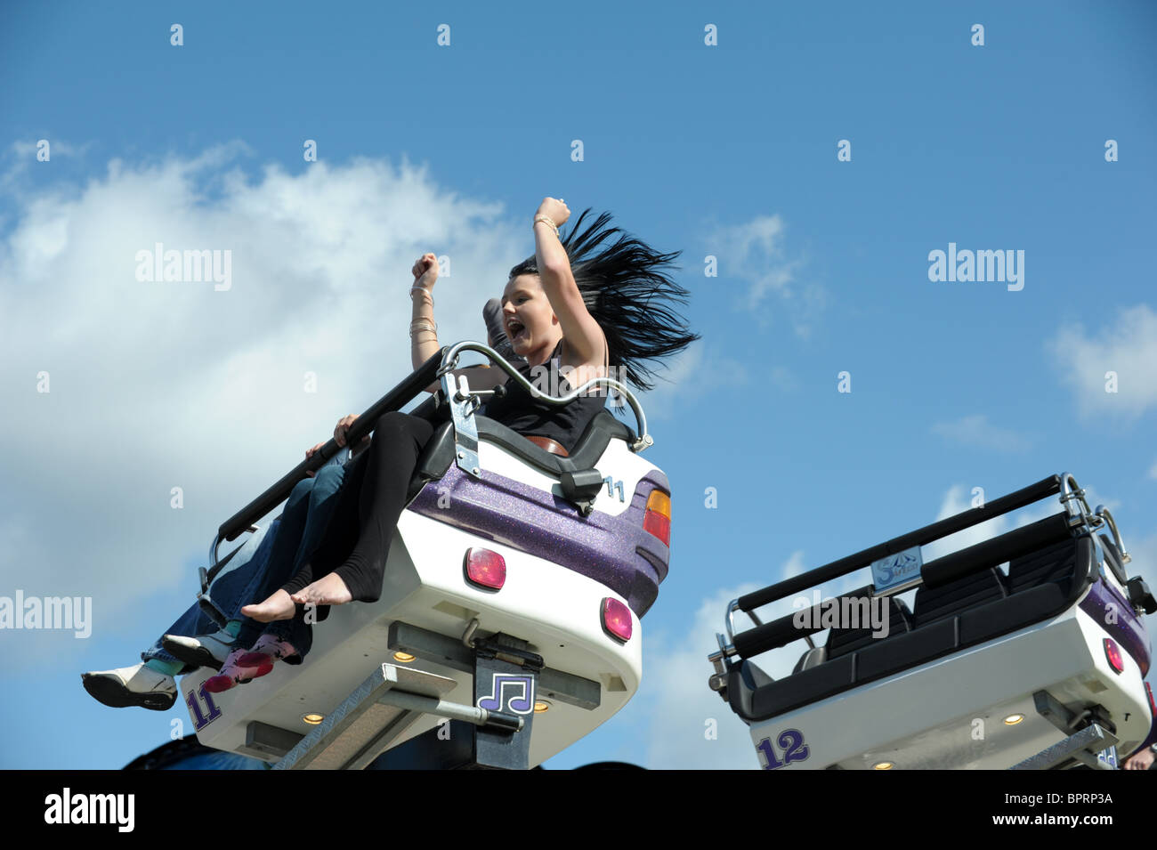 Girls laughing and screaming on a fairground ride with arms in the air and black hair flying up against a blue sky Stock Photo