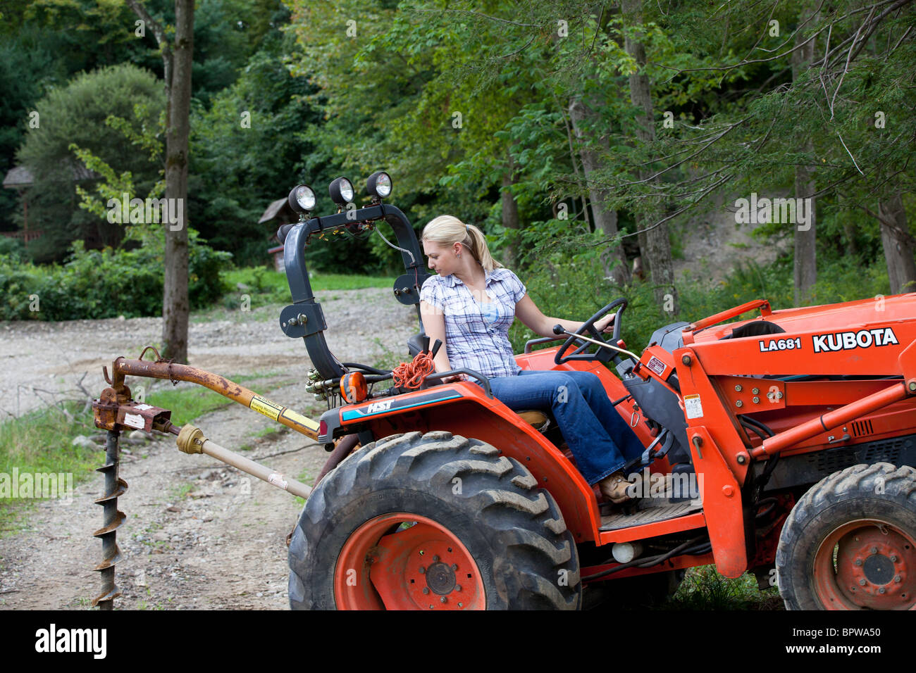 Young blond woman driving Kubota tractor with post hole digger attachment working on farm Stock Photo