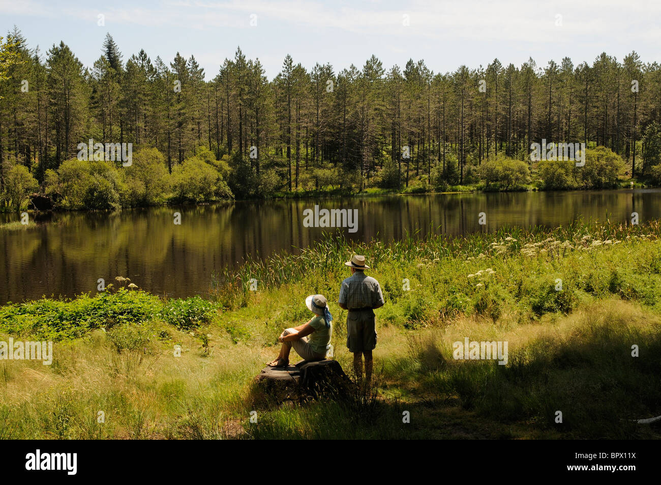 Couple sitting at the waters edge Lake and pine forest situated on a plateau in the mountains de l' Epinouse southern France Stock Photo