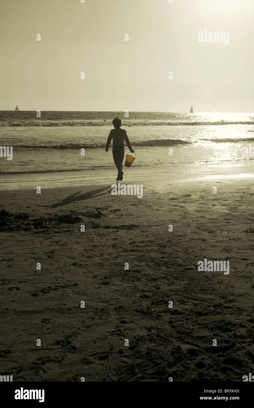 sepia image of boy running on beach with bucket near the ocean Stock Photo