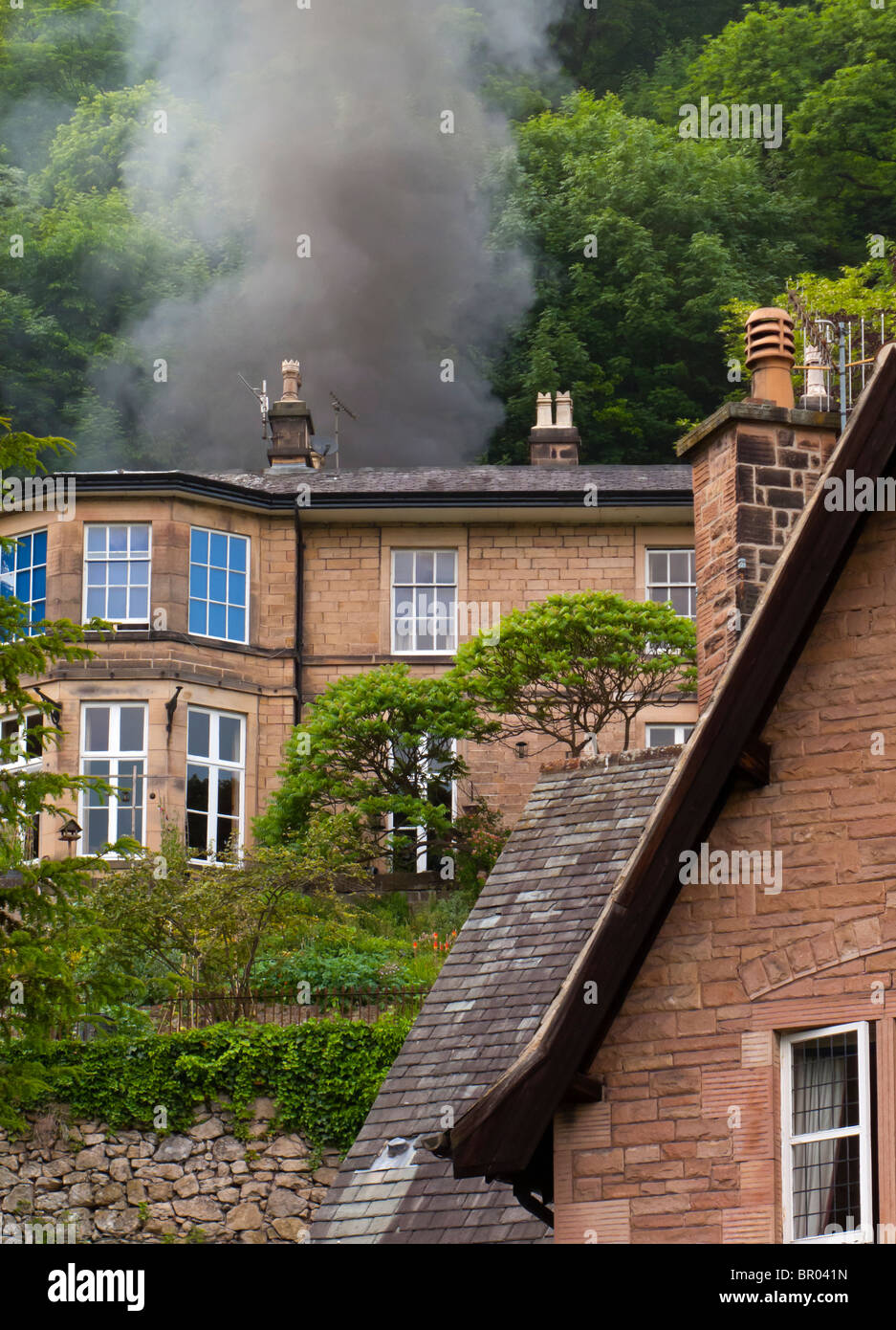 House on fire with black smoke billowing from the roof and trees in background Stock Photo