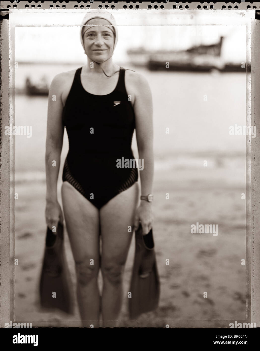Middle Aged Caucasian woman swimmer on a beach wearing a dark swimming suit holding flippers in each hand. Stock Photo