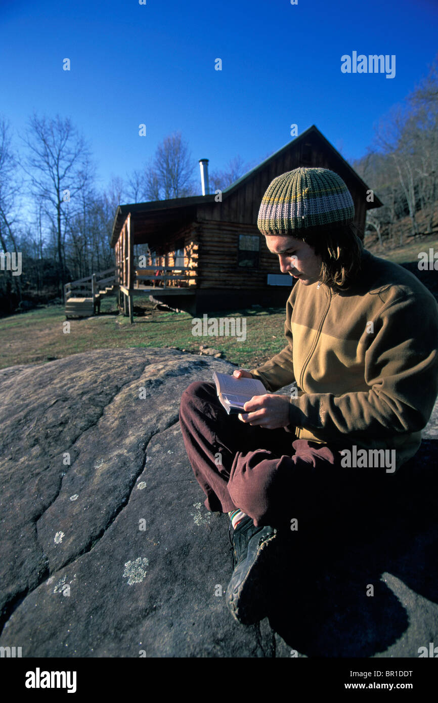 A Male Climber Reading A Book On A Rock By A Cabin In The Woods
