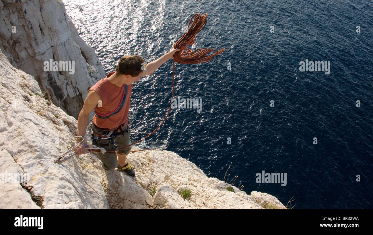 Rockclimber wearing an orange tanktop throws a rope over cliff towards the ocean. Stock Photo