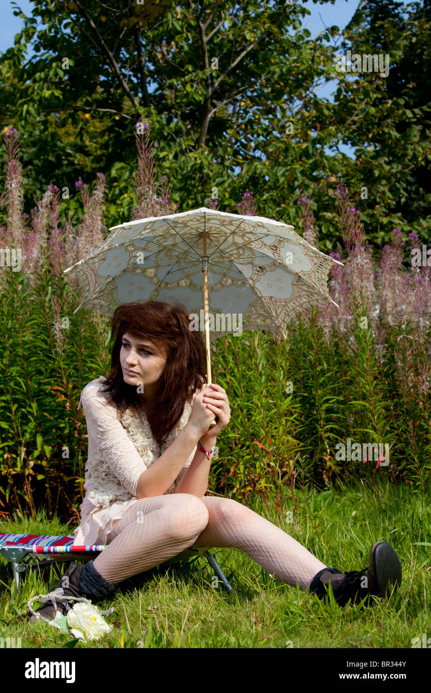 A model with long dark hair and wearing light coloured clothing poses in an overgrown garden with a parasol. Stock Photo