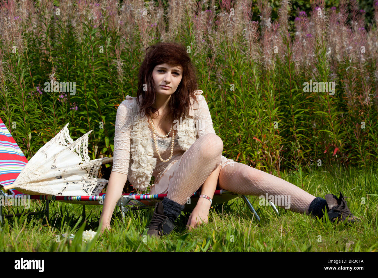 A model with long dark hair and wearing light coloured clothing poses in an overgrown garden with a parasol. Stock Photo
