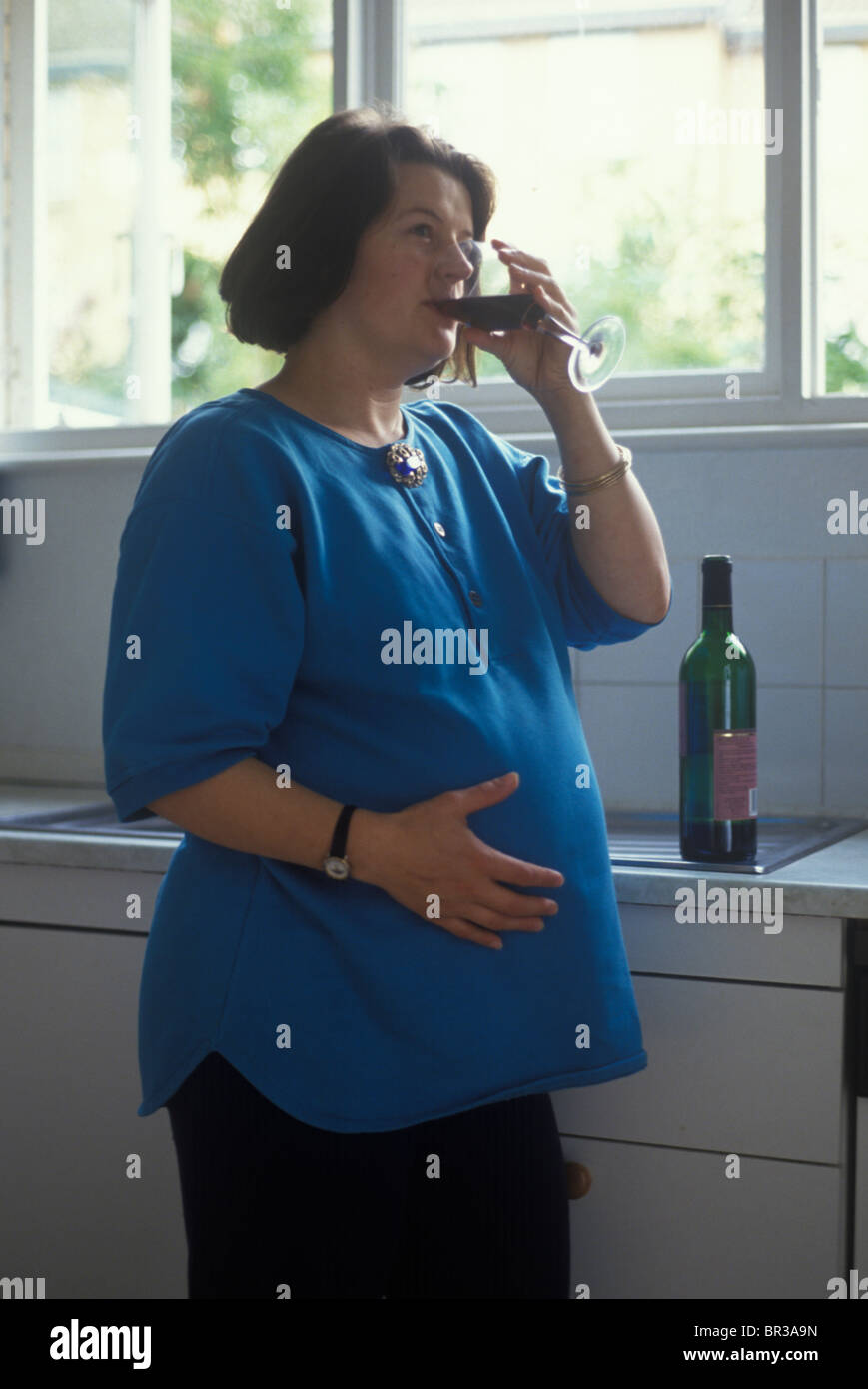 heavily pregnant woman drinking a glass of red wine in her kitchen