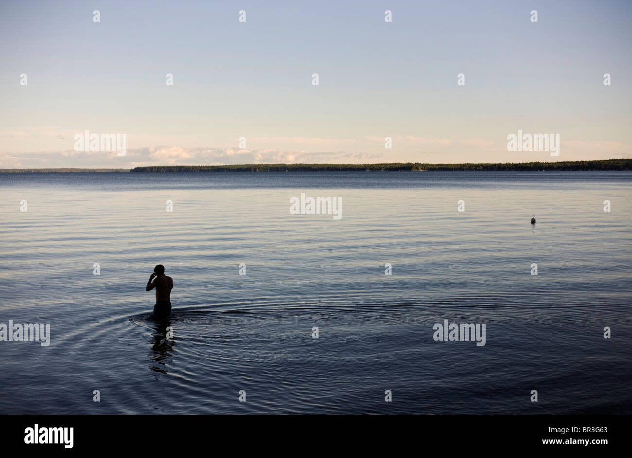 A man, silhouetted agains the calm water, wades into dark, blue water at dusk. Stock Photo
