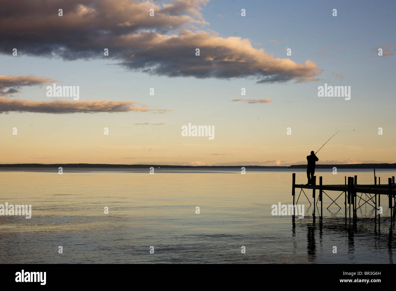A man casts his fishing line from a silhouetted dock above the calm, clear water. Stock Photo
