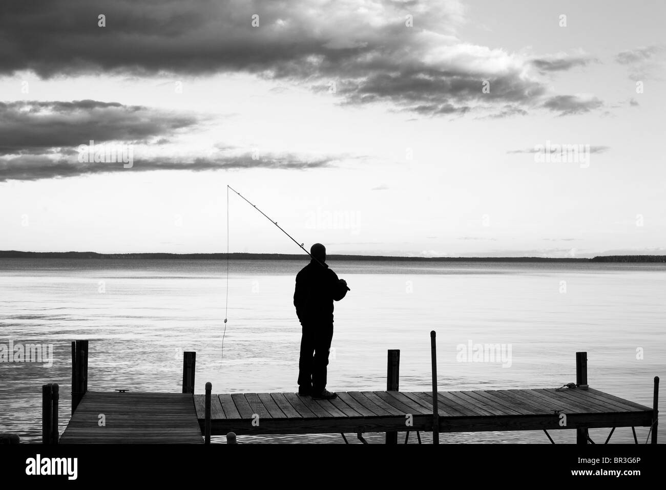 A black and white image of a man, silhouetted against a dramatic sky, holding a fishing rod. Stock Photo