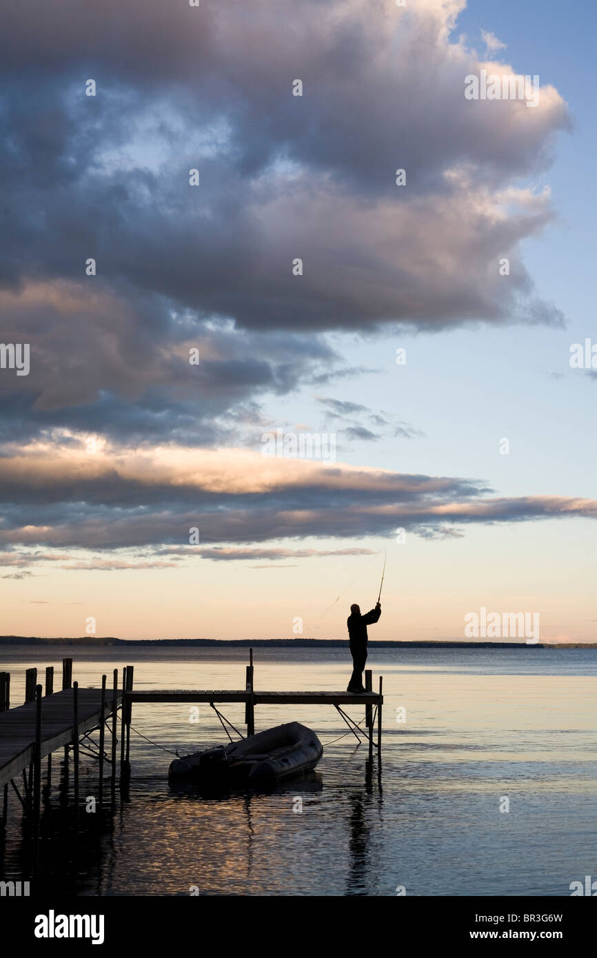 A man silhouetted against a dramatic sky at dusk, casts his fishing line into the calm, clear, smooth waters Stock Photo