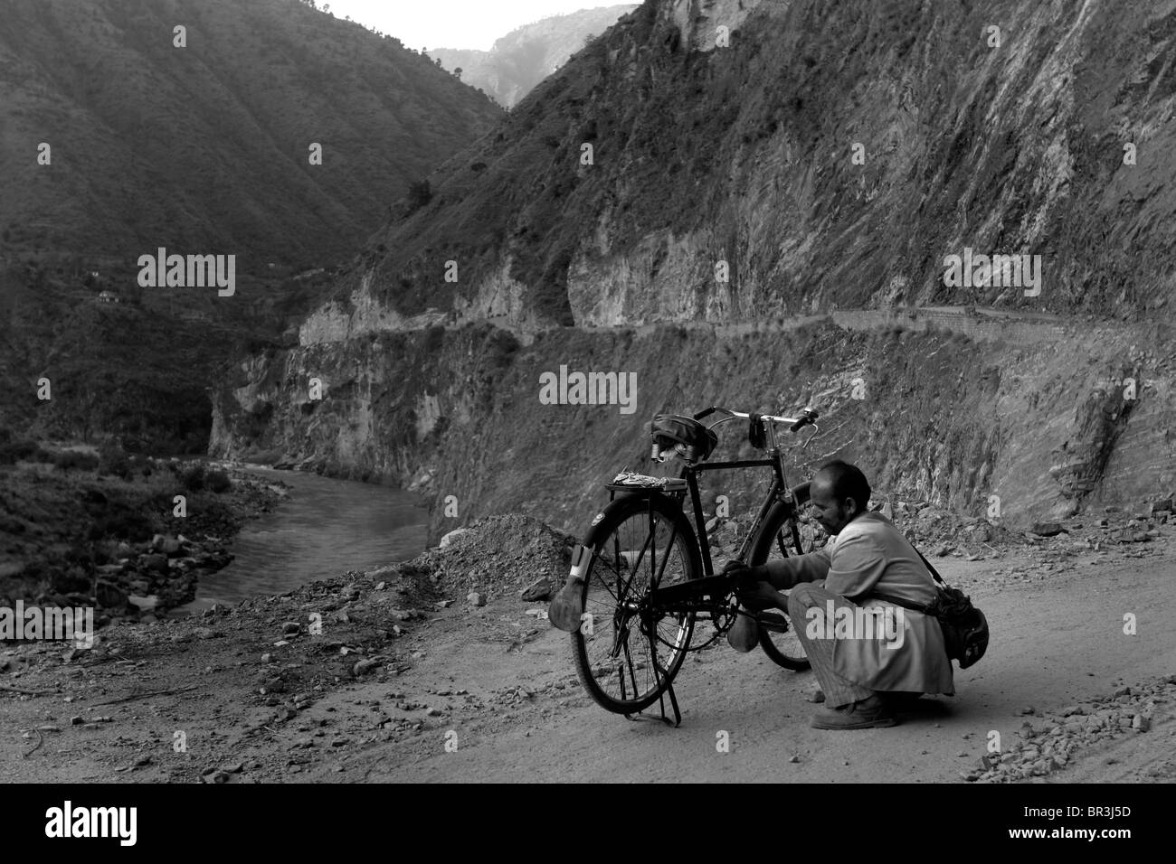 A man repairs a flat punctured tire on his bicycle on a remote mountain road in the Himalayas in Himachal Pradesh in India. Stock Photo