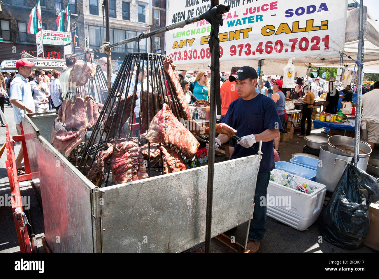 young Hispanic man tends rotating rack roasting meat as big crowd wanders 9th Avenue at annual International food festival Stock Photo