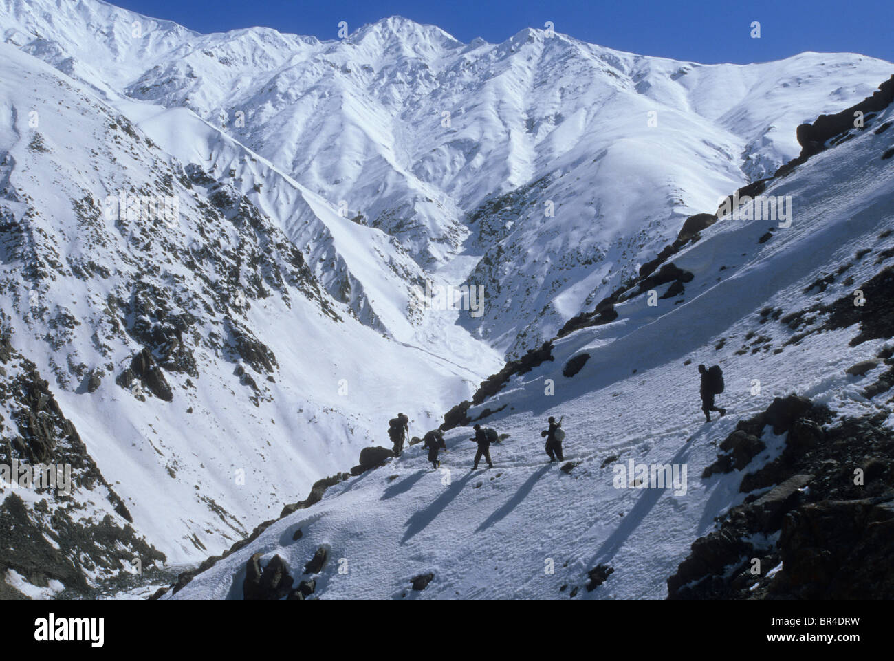 Men of the Wakhi tribe hike along a snow covered trail above the Wakhan River below peaks of the Hindu Kush mountains, Wakhan Co Stock Photo
