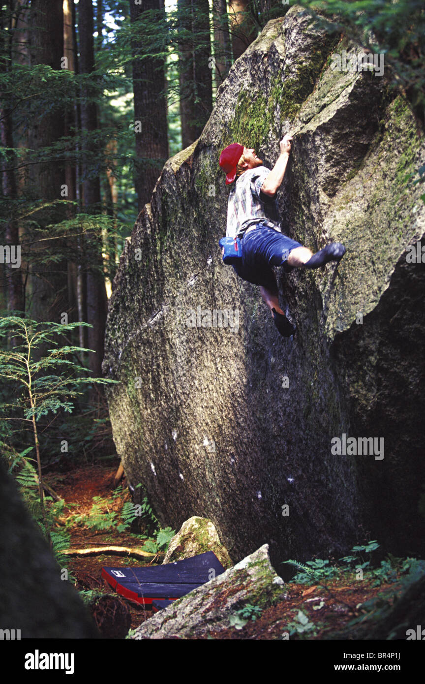Man wearing a red hat and shorts clings to a granite boulder in the forest. Stock Photo