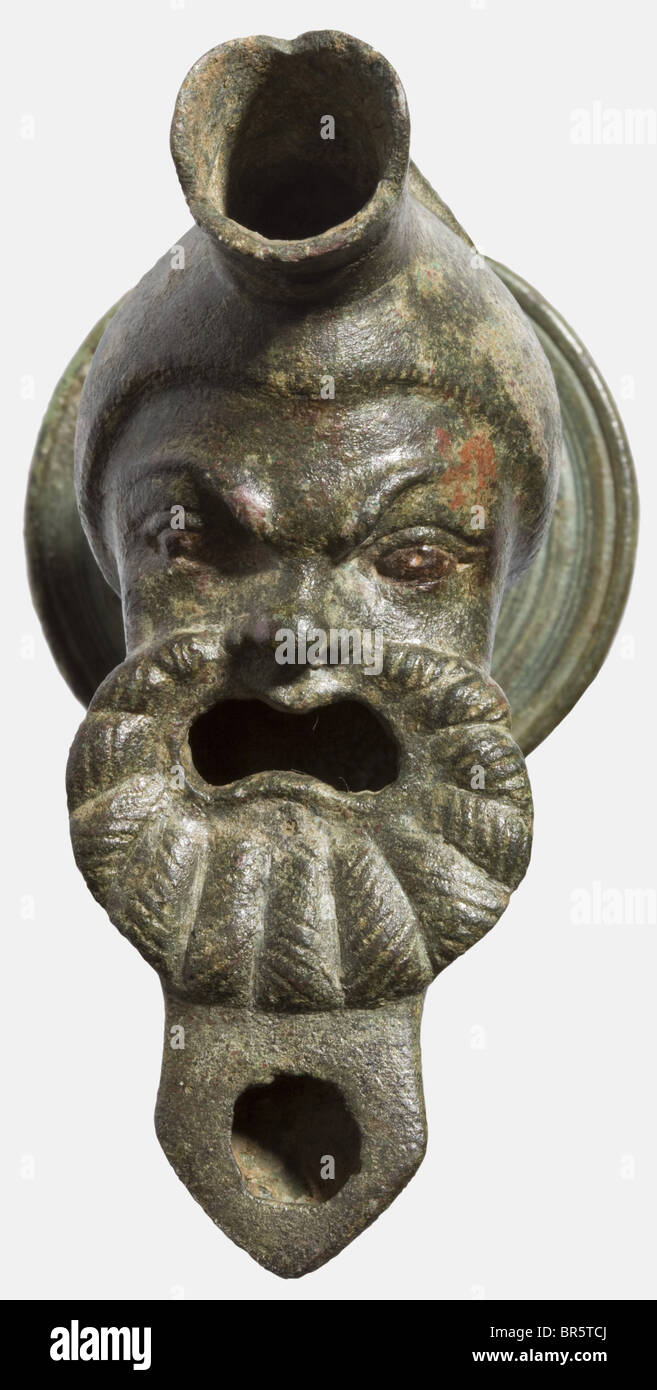 A Roman bronze lamp with tripod stand, 1st/2nd century A.D. Cast bronze complemented by cold-working. Made of two pieces. The lamp in the shape of a bearded comedian's mask, the mouth - which was filled with oil - wide open, expressive eyes, the brows and nose distinctively shaped. Incl. a tripod stand terminating in three stylised, flared animal legs with accurately fitting mounting. Finely chiselled. Height 16.8 cm. Slightly spotted patina, small chipping at the front of the wick hole, small repair at the stem. Cf. Peter La Baume, Römisches Kunstgewerbe, 1983, Stock Photo