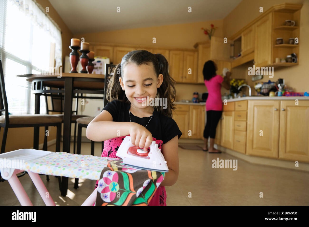 Girl using toy iron and ironing board in kitchen Stock Photo