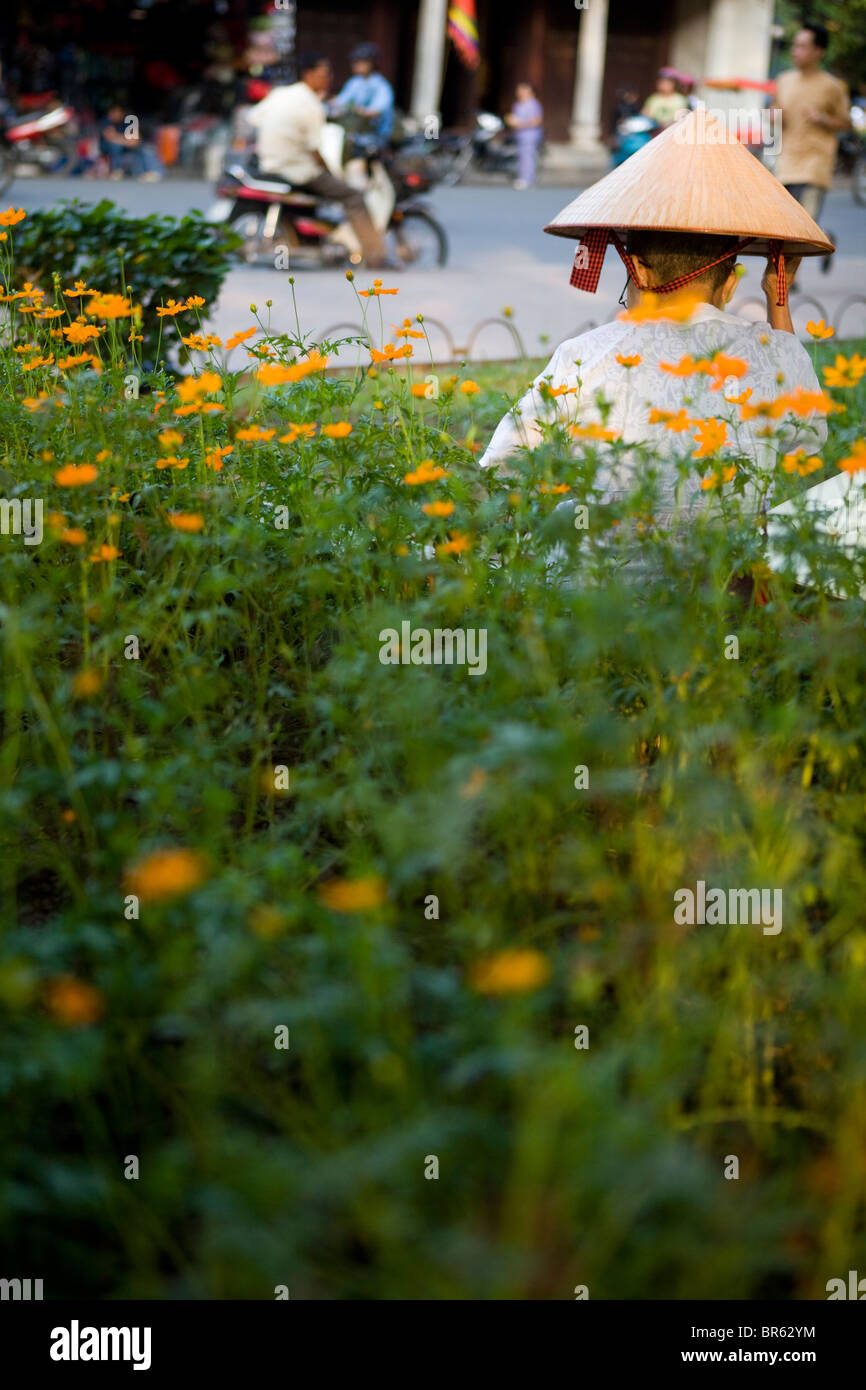 Woman with conical hat sitting amongst flowers in busy Hanoi street, Vietnam Stock Photo