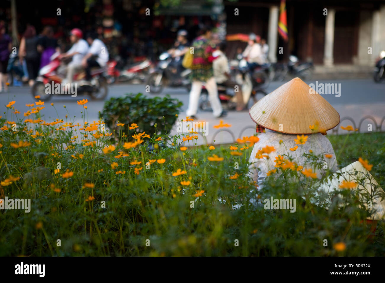 Woman with conical hat sitting amongst flowers in busy Hanoi street, Vietnam Stock Photo