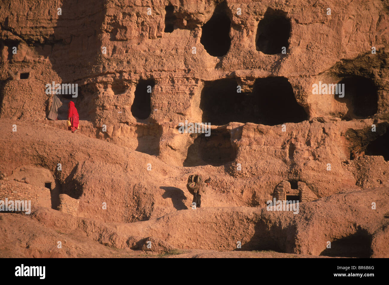 Women carry water past caves carved into a cliff Bamiyan Valley Stock Photo
