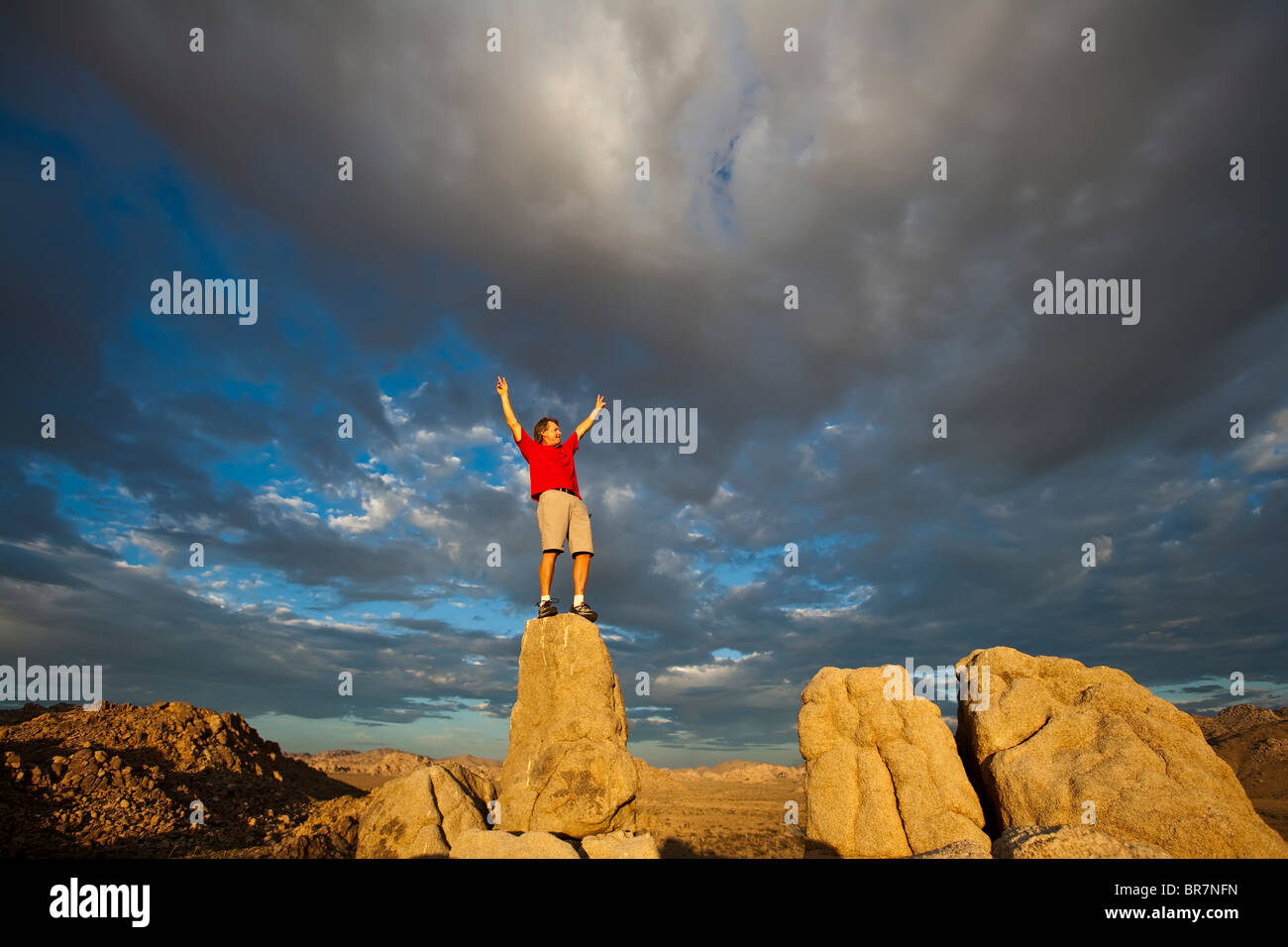 Rock climber leaps across a gap  on the summit of a pinnacle after a successful ascent as storm clouds build in the setting sun. Stock Photo