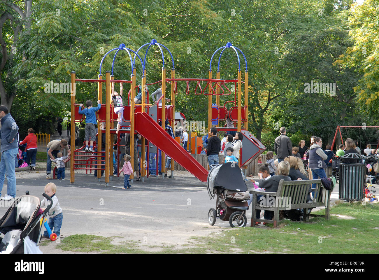 Families and children play in a leafy park playground on a sunny day