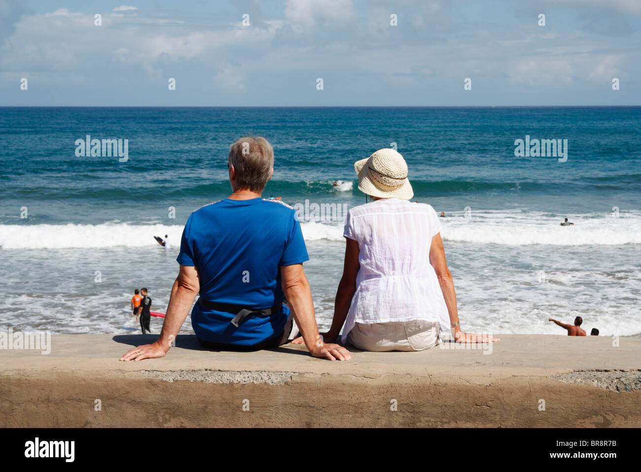Couple sitting on sea wall watching surfers in Spain Stock Photo - Alamy