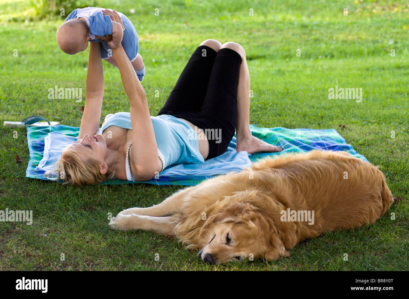 Mother playing with her baby in the park with golden retriever lying down beside. Stock Photo