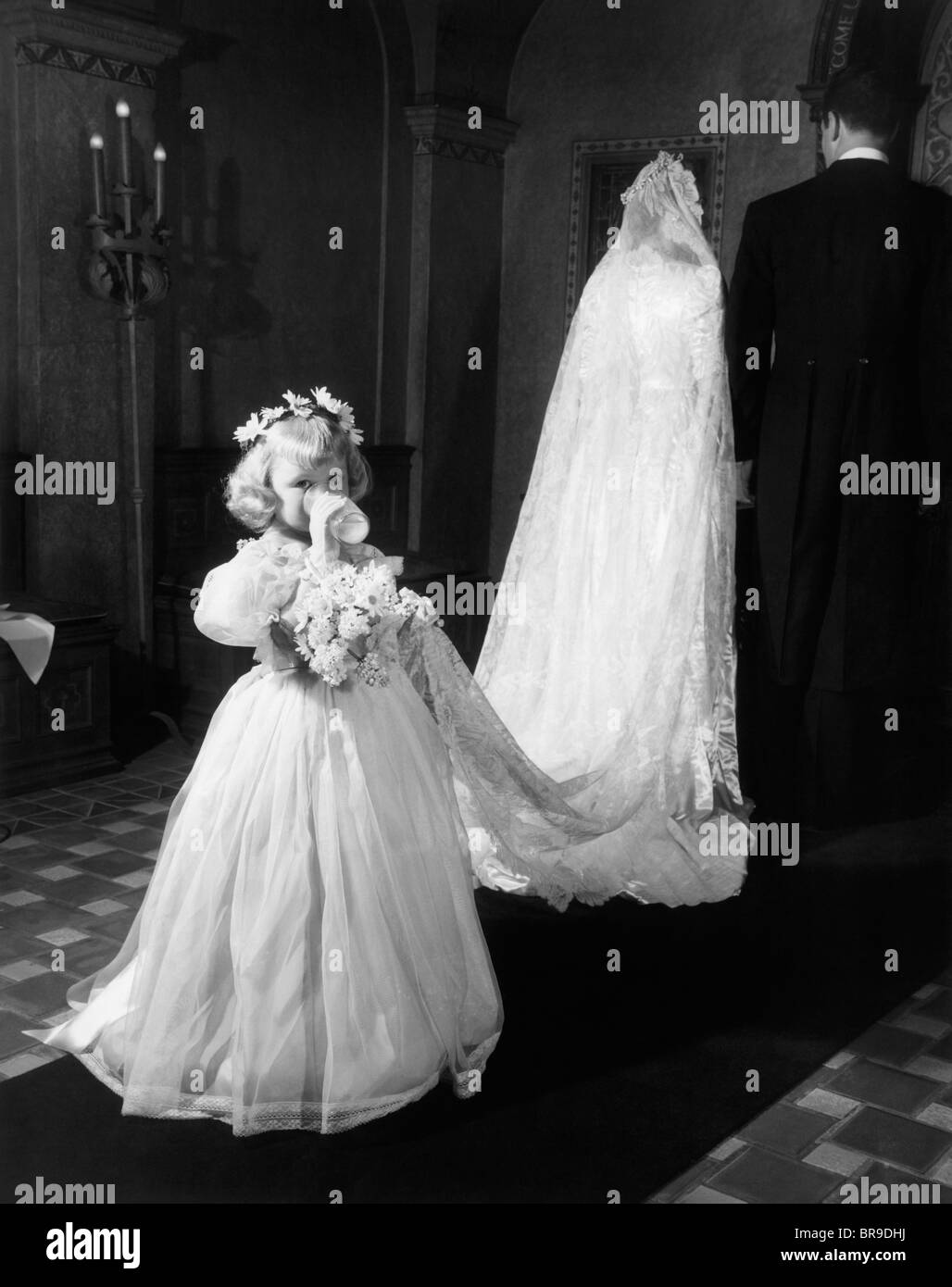 1950s LITTLE GIRL BRIDESMAID DRINKING GLASS OF MILK HOLDING BRIDAL VEIL TRAIN WALKING DOWN CHURCH AISLE LOOKING AT CAMERA Stock Photo