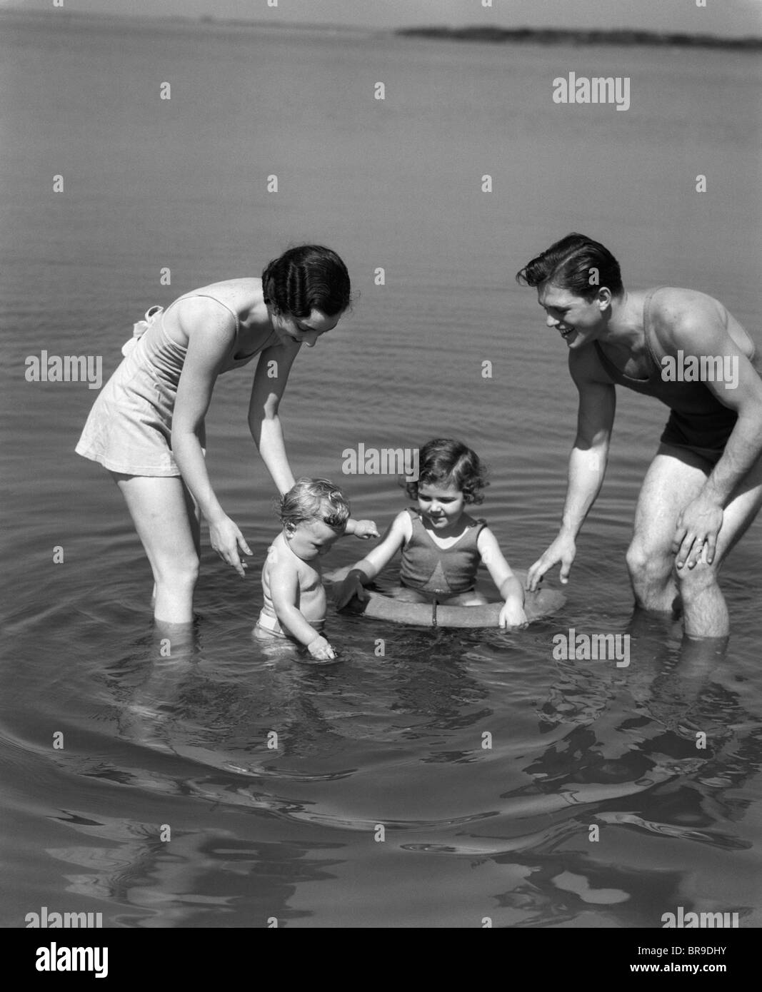 1930s FAMILY FATHER MOTHER DAUGHTER SON WITH RUBBER INNER TUBE WADING IN SEASHORE WATER Stock Photo