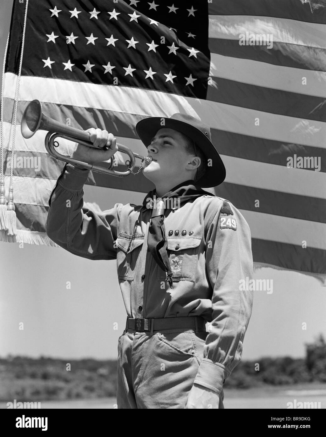 1940s BOY SCOUT PLAYING BUGLE IN FRONT OF 48 STAR AMERICAN FLAG Stock
