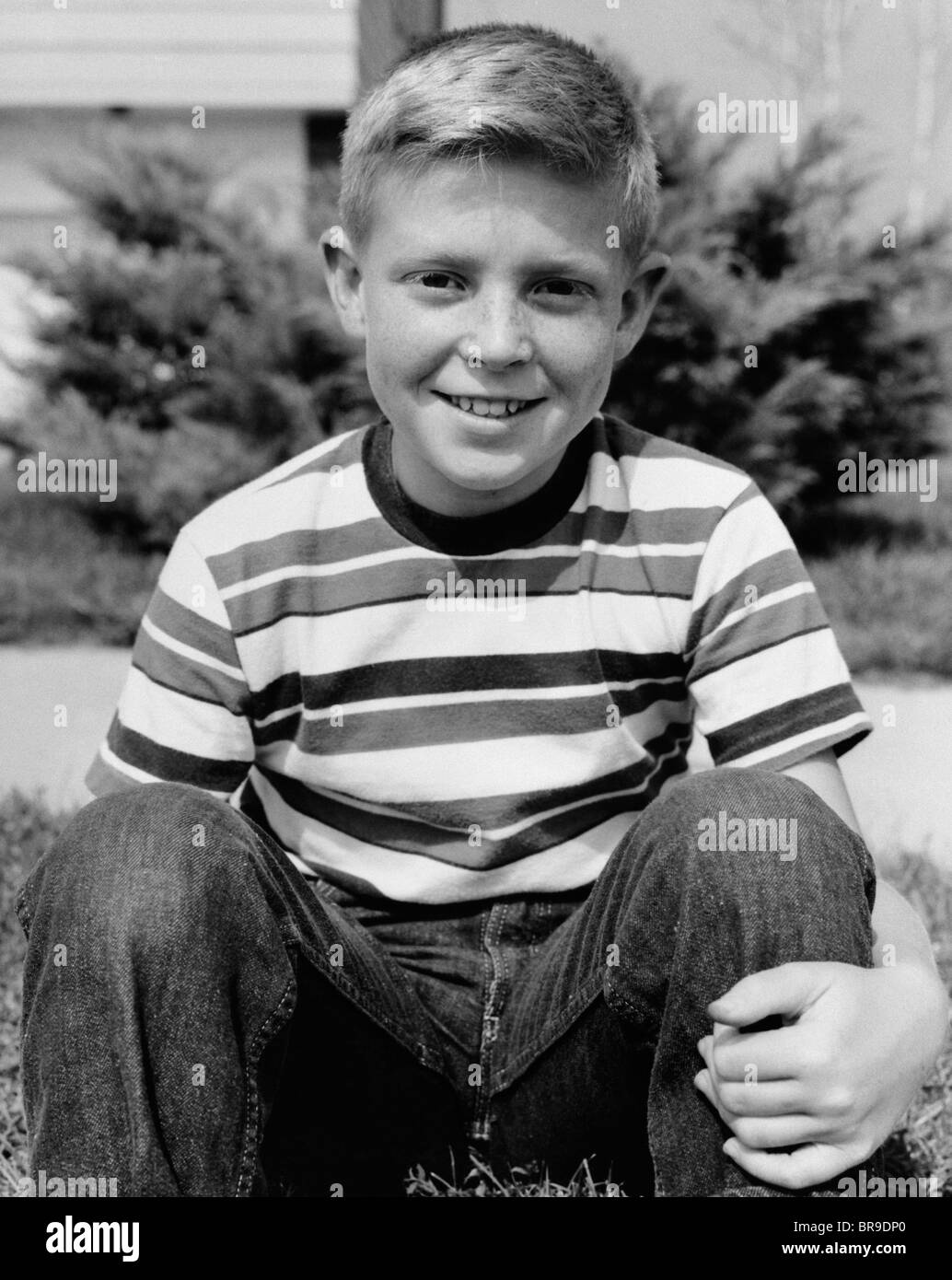 1960s SMILING BOY SITTING WEARING BLUE JEANS AND STRIPED JERSEY SHIRT LOOKING AT CAMERA Stock Photo