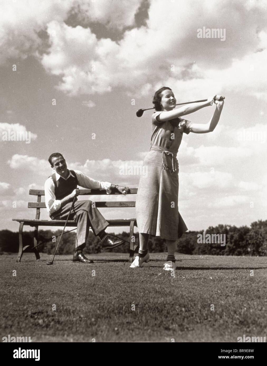 1930s COUPLE PLAYING GOLF MAN ON BENCH WOMAN SWINGING CLUB Stock Photo