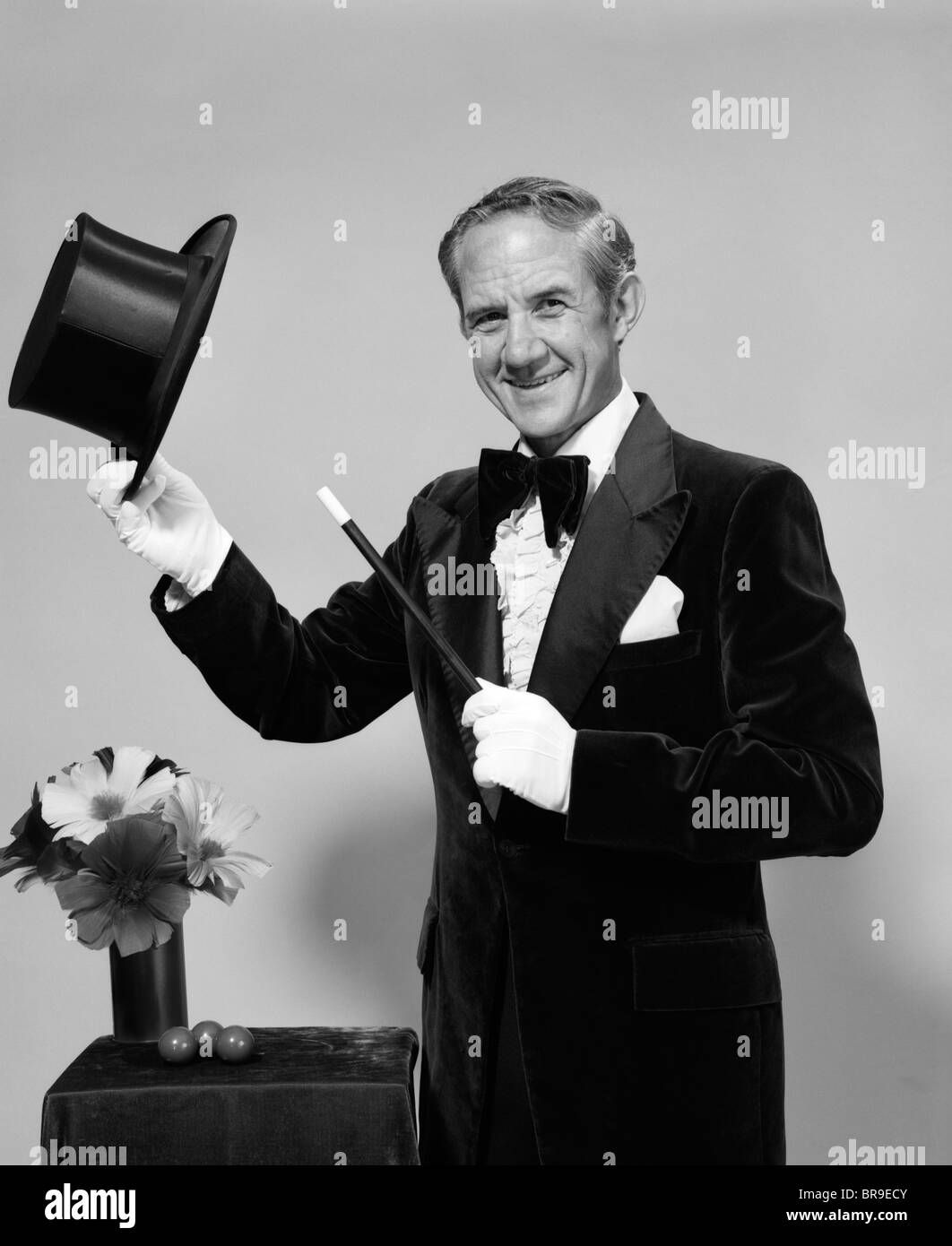 1970s SMILING MAN MAGICIAN WEARING VELVET TUXEDO WHITE GLOVES POINTING MAGIC WAND AT TOP HAT LOOKING AT CAMERA Stock Photo