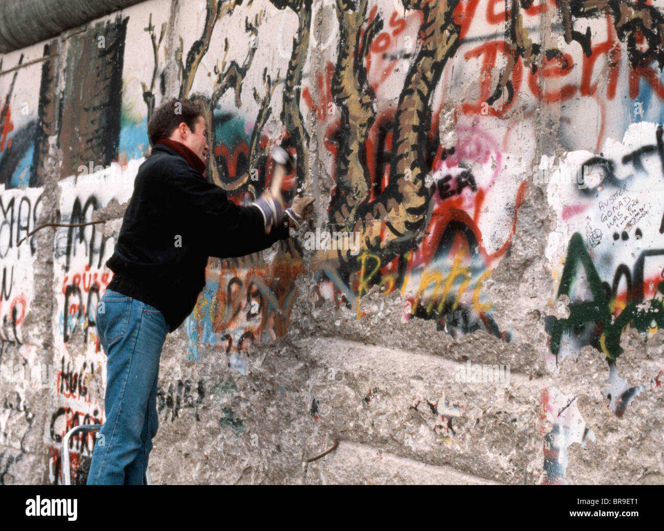 1980s TEENAGE BOY CHIPPING AWAY AT THE GRAFFITI SPRAY PAINTED BERLIN WALL WITH HAMMER AND CHISEL NOVEMBER 1989 Stock Photo