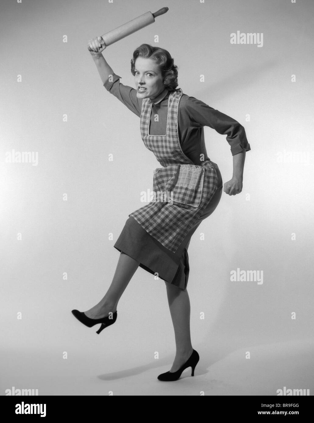 1950s ANGRY HOUSEWIFE IN APRON WIELDING ROLLING PIN OVER HER HEAD Stock Photo