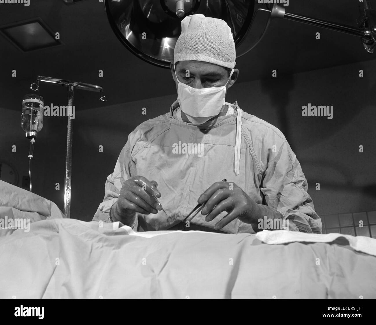 1950s SURGEON STANDING INSIDE OPERATING ROOM ABOUT TO OPERATE ON PATIENT Stock Photo