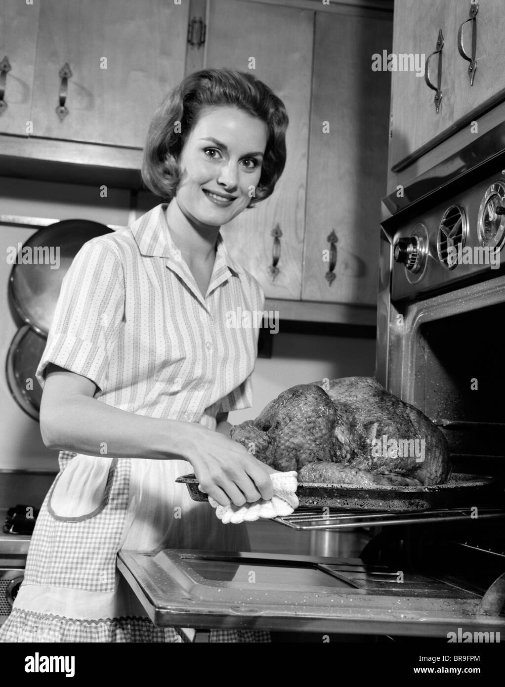 1960s SMILING HOUSEWIFE REMOVING THANKSGIVING ROAST TURKEY FROM OVEN LOOKING AT CAMERA Stock Photo